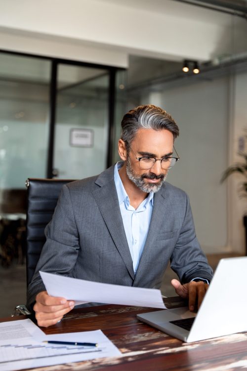 usy Latin or Indian male business man accountant analyst holding documents, work at laptop computer doing online trade market tech research. Focused Hispanic businessman with paperwork in office.