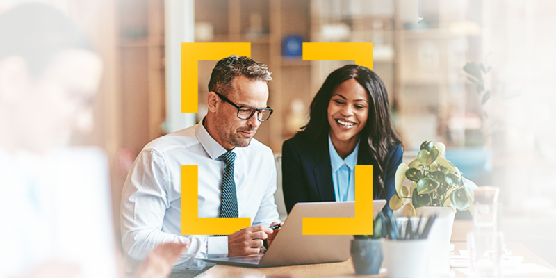 Two coworkers collaborating at a desk with a tablet and laptop in a bright office setting.