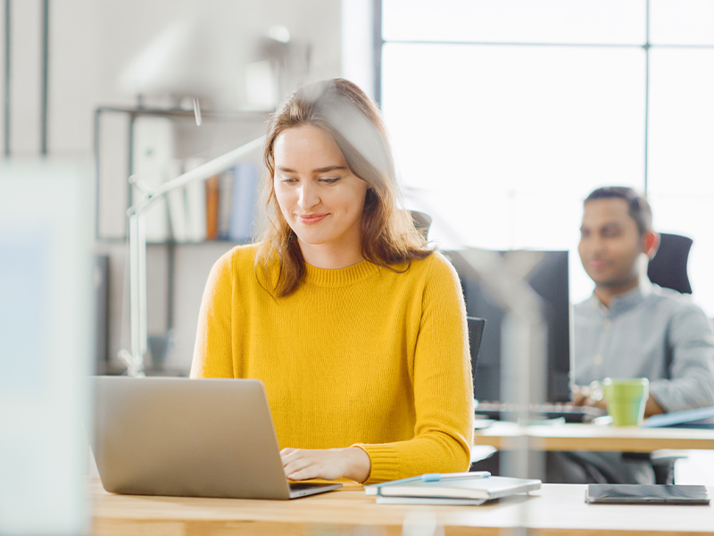 Woman in a mustard sweater types on a laptop at a bright office desk, others working in the background.