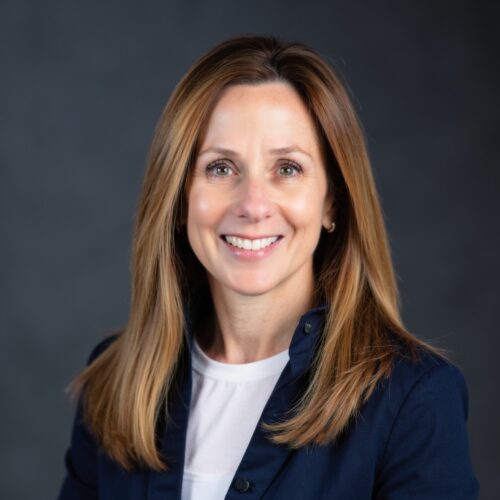 Smiling woman portrait with long light brown hair, wearing a navy blazer and white top, against a dark background (professional headshot).