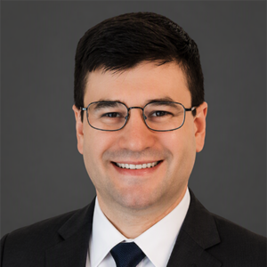Professional headshot of a smiling man in a dark suit and glasses against a gray backdrop.
