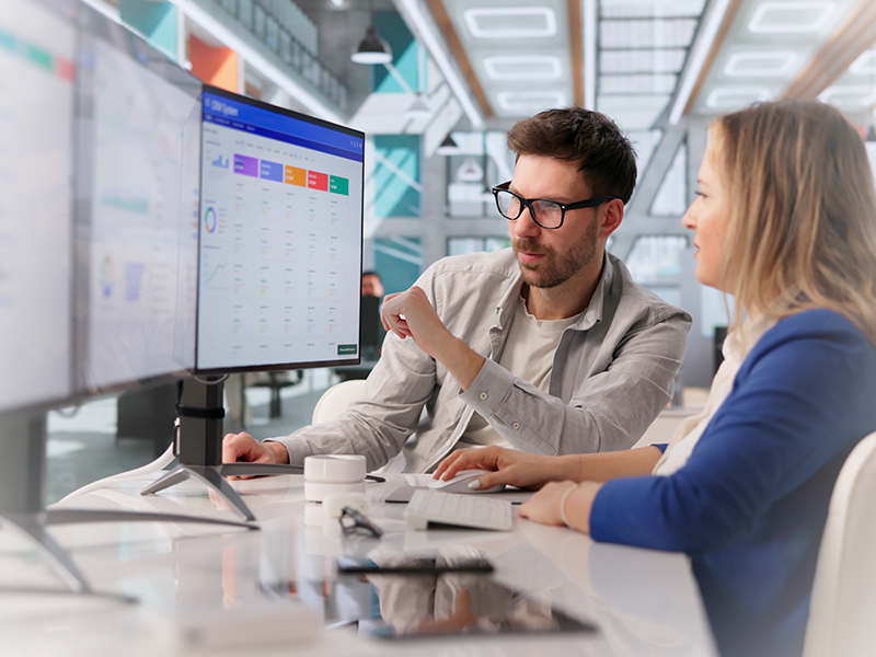 Two colleagues review a data dashboard on a large monitor in a bright, modern office.