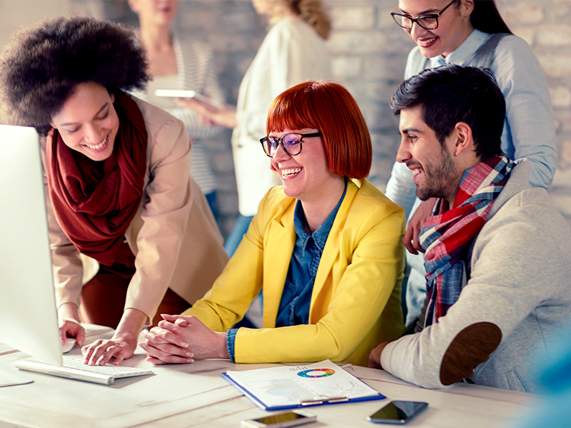 A group of coworkers gathered around a desktop computer, smiling and collaborating in a bright office setting.