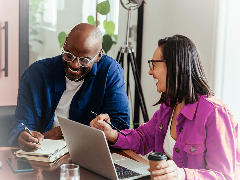 Two coworkers collaborate at a desk: a man writes in a notebook while a woman points at a laptop and smiles.