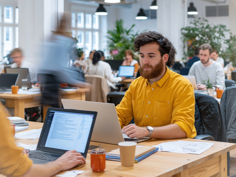 Man with a mustard-yellow shirt typing on a laptop in a bright open-plan workspace with colleagues in the background.