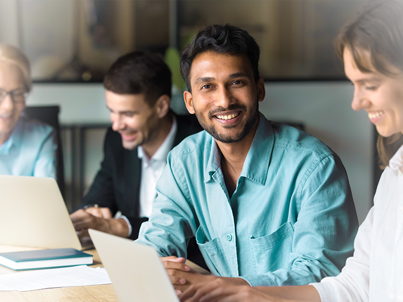A group of coworkers smiling and working at a table with laptops in a bright office.