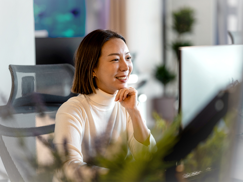 Woman in a white turtleneck smiling while working at a desk with a computer monitor in a bright office.