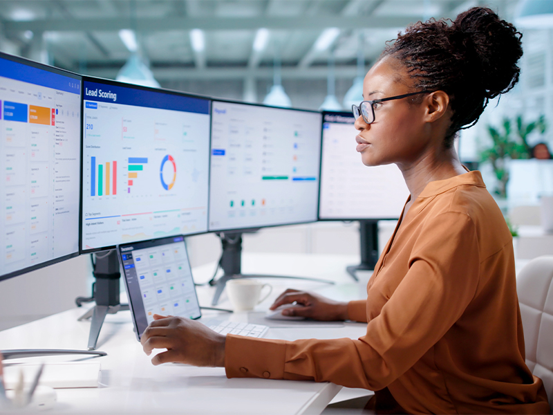 Woman analyzing data dashboards on a multi-monitor setup in a modern office. Her tablet is on a stand while she types on a keyboard.