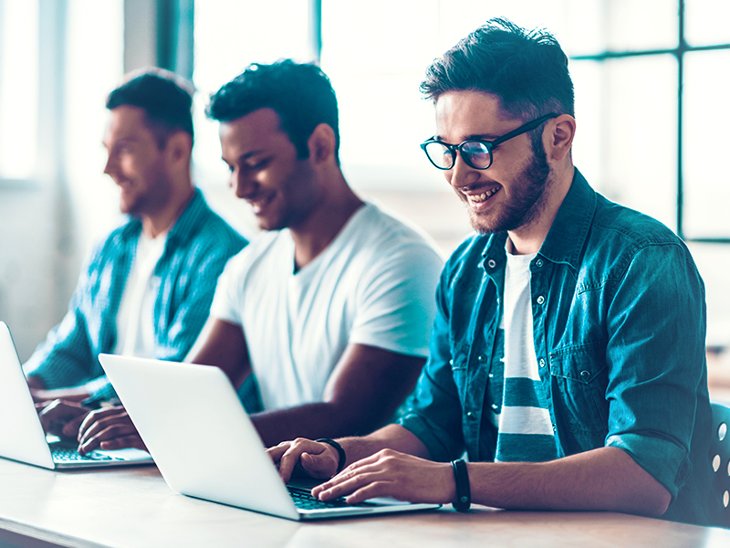Three coworkers working on laptops at a bright office desk, smiling.
