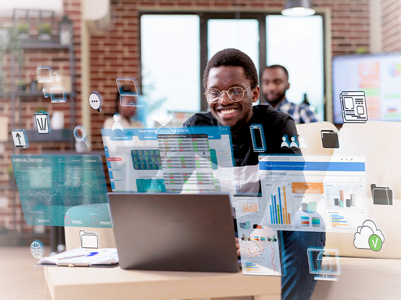 Smiling man at a laptop surrounded by floating data charts and icons in a bright office setting.