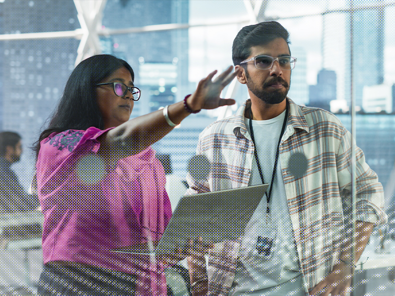 Two coworkers collaborate in a bright office, one gesturing toward a glass wall while the other holds a laptop and listens.
