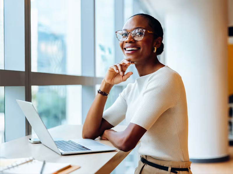Smiling woman wearing glasses sits at a desk with an open laptop by a large window in a bright office environment.