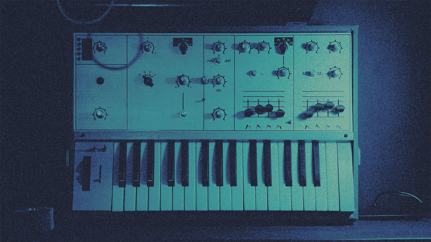 Vintage analog synthesizer with numerous knobs and a piano-style keyboard in a dimly lit room in a blue tint.
