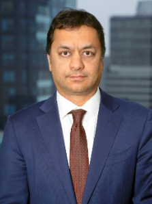 Professional headshot of a man in a navy suit and brown tie against a blurred office backdrop.