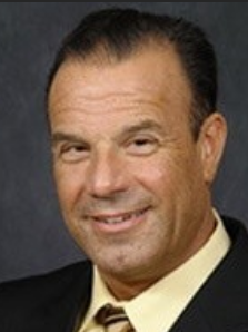 Head-and-shoulders portrait of a smiling middle-aged man in a dark suit and yellow striped tie against a gray backdrop.