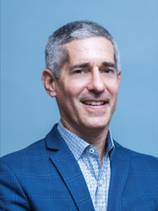 Professional headshot of a smiling man in a blue blazer and light blue checkered shirt against a blue background.