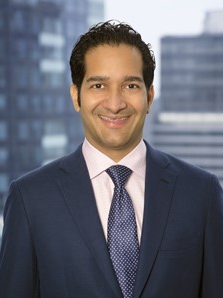 Professional headshot of a smiling man in a navy suit, pink shirt, and purple tie, with a blurred cityscape background.