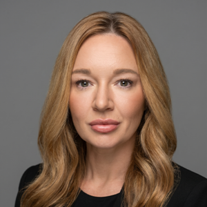 Professional headshot of a woman with long blonde hair wearing a black blazer against a gray background.