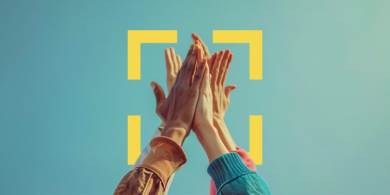 Diverse hands coming together in a high-five against a blue sky, with yellow corner-bracket graphics framing the scene.
