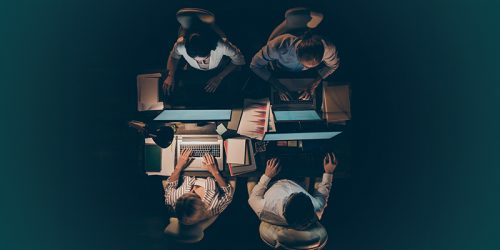 Top-down view of four people around a table working on laptops with documents and notebooks_autogenerated.