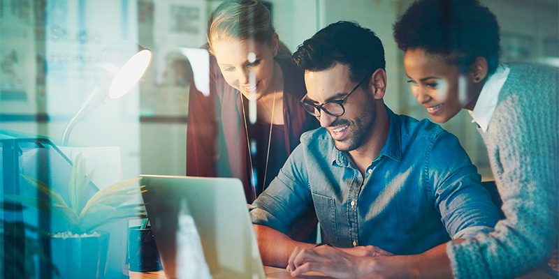 Three coworkers gathered around a laptop, smiling and collaborating at a desk in a modern office.
