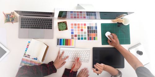 Top-down view of a design workspace with a laptop, color swatches, keyboard, tablet, mouse, and cutting mat as hands work on projects.