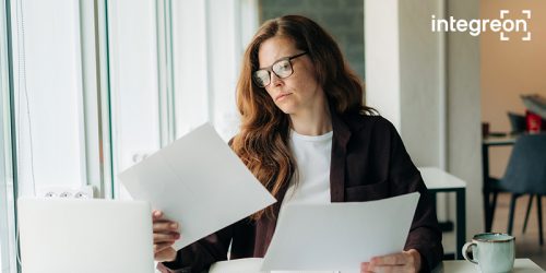 Professional woman with glasses reviewing documents at a sunlit office desk by a window, logo in corner.