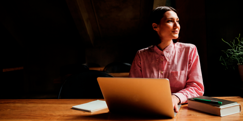 Woman in a pink shirt sits at a wooden desk with an open laptop, a notebook, and a stack of books, gazing to the right in a dimly lit room with a plant in the background.