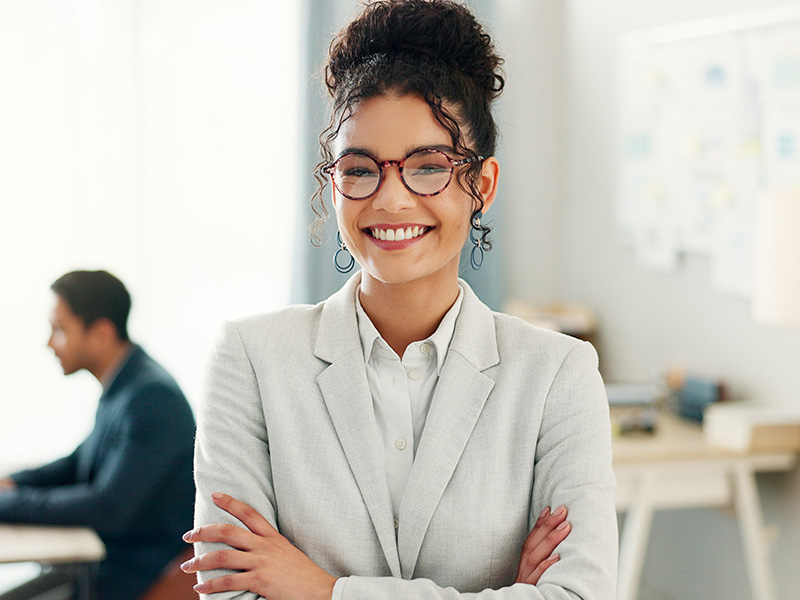 Smiling woman with curly hair and glasses in a light gray blazer stands in an bright office, with a blurred colleague in the background.