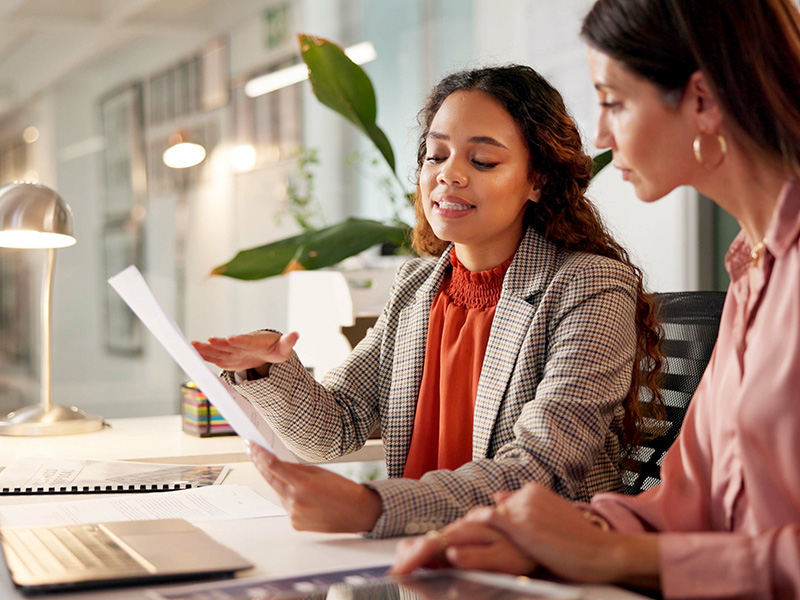 Two women collaborate at a desk, one pointing to a document as the other looks on in a bright office with a laptop nearby.