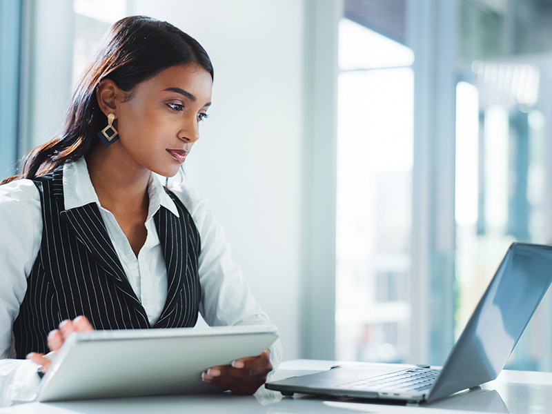 Businesswoman working on a tablet at a desk with a laptop in a bright office setting.