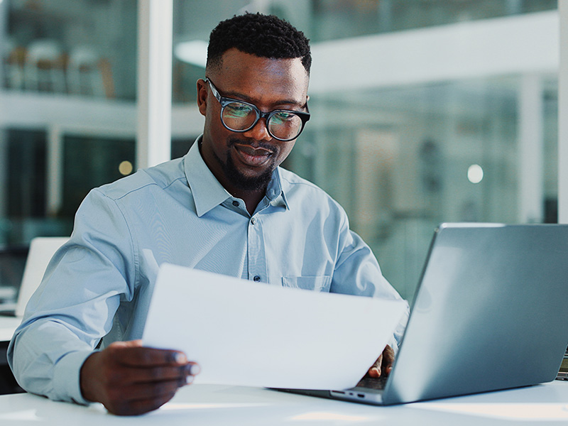 Man in glasses reads a document at a desk with a laptop in an office setting.