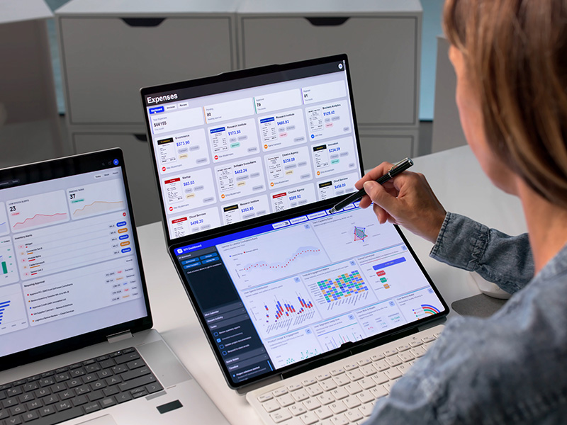 Person using a foldable laptop to review an expense dashboard on the top screen and charts on the bottom screen, with a stylus in hand.