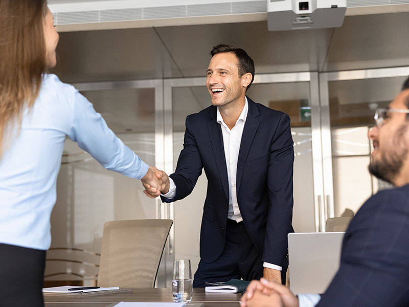 Man in a navy suit shakes hands with a woman across a conference table, smiling in a bright office environment.