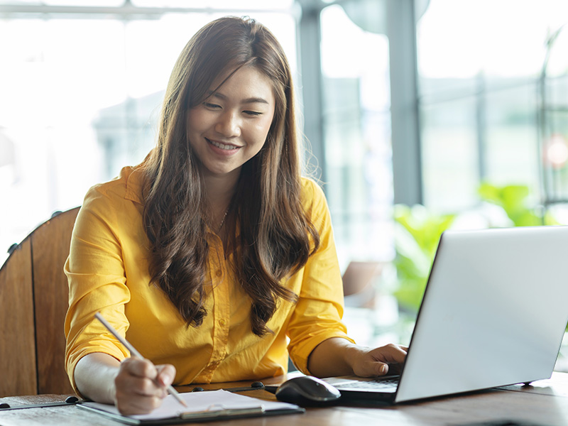 Smiling woman in a yellow shirt writes on a notebook at a desk with a laptop nearby in a bright office setting.