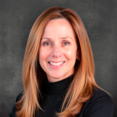 Portrait of a smiling woman with long light brown hair wearing a black turtleneck, against a gray studio backdrop.