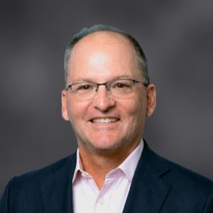 Professional headshot of a smiling middle-aged man with short gray hair and glasses, wearing a pink shirt and navy blazer.