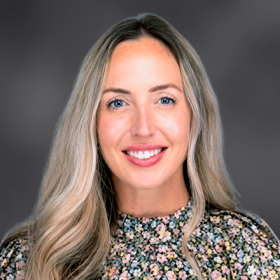 Smiling woman with long blonde hair wearing a floral blouse, posed against a dark blurred background.
