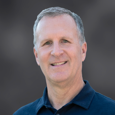 Portrait of a smiling, middle-aged man with short gray hair wearing a navy polo shirt against a blurred gray background.