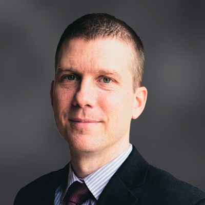 Head-and-shoulders portrait of a man in a dark blazer, striped shirt, and red tie, against a gray gradient background.