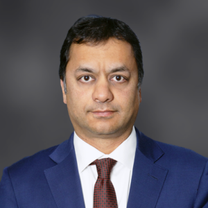 Professional headshot of a man in a navy suit and red tie, looking at the camera against a gray backdrop.