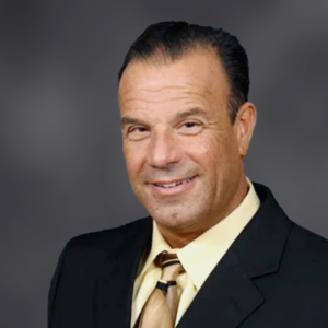 Professional headshot of a smiling man in a black suit and tan tie against a grey backdrop.