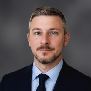 Professional head-and-shoulders portrait of a man in a navy suit and blue tie against a neutral gray backdrop.