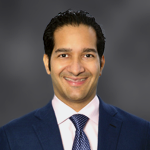 Portrait of a smiling man wearing a navy suit, white shirt, and patterned tie against a gray gradient background (professional headshot).
