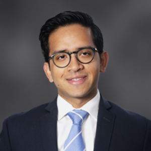 Professional headshot of a man wearing a dark suit, white shirt, and light blue striped tie, smiling against a neutral gray background.